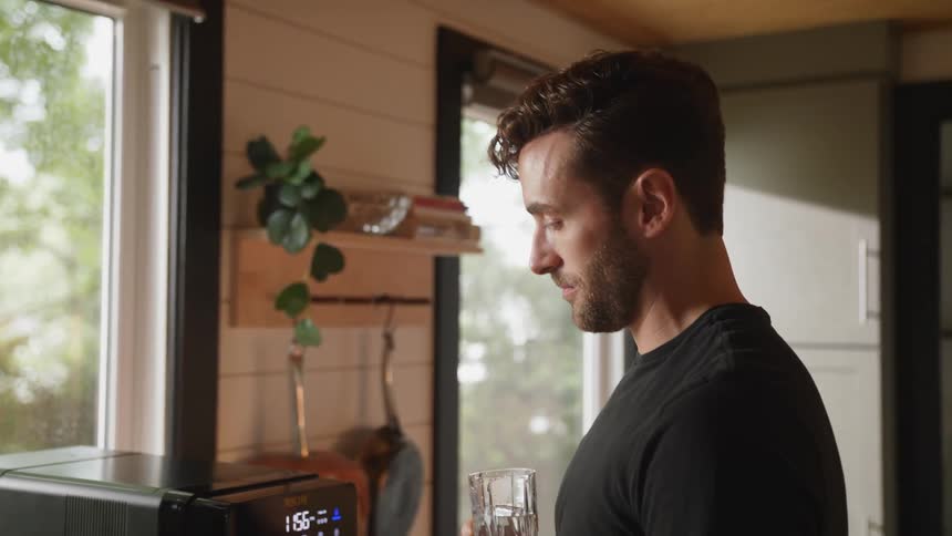 Man with beard and black t shirt drinks a glass of water in the kitchen