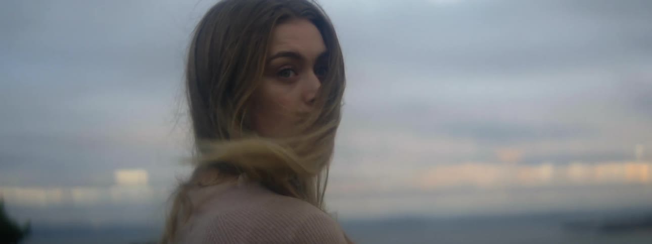 Blonde woman wearing brown sweater looks over her shoulder in the field at dusk