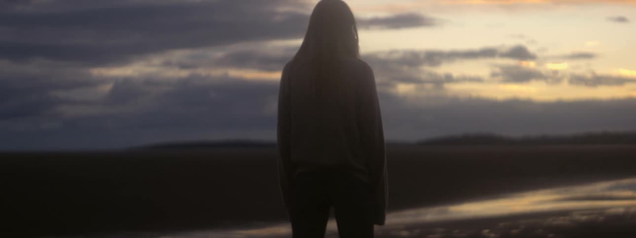 A woman stands motionless and stares at a river in front of her