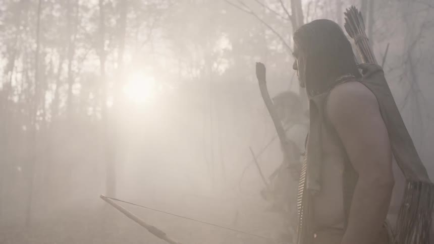 Native americans carrying weapons quietly walk through a misty forest