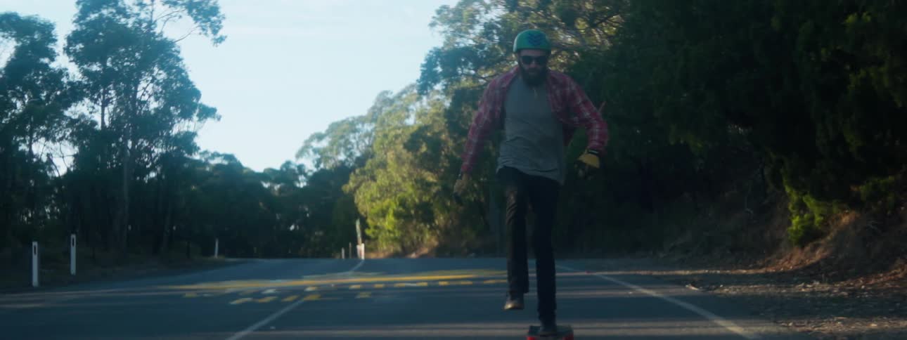 Man wearing turquoise helmet red shirt and black pants skateboards on country road