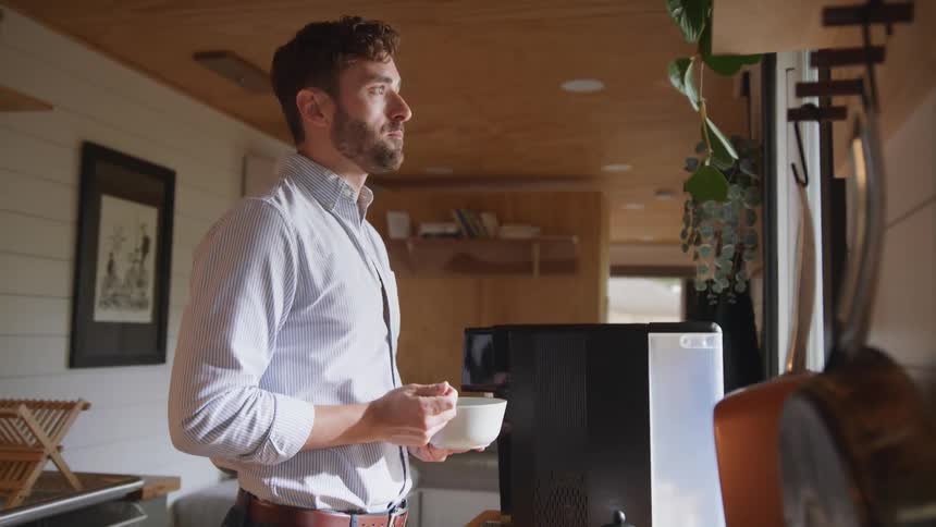 A man stands at a small table in his kitchen while eating food from a glass bowl