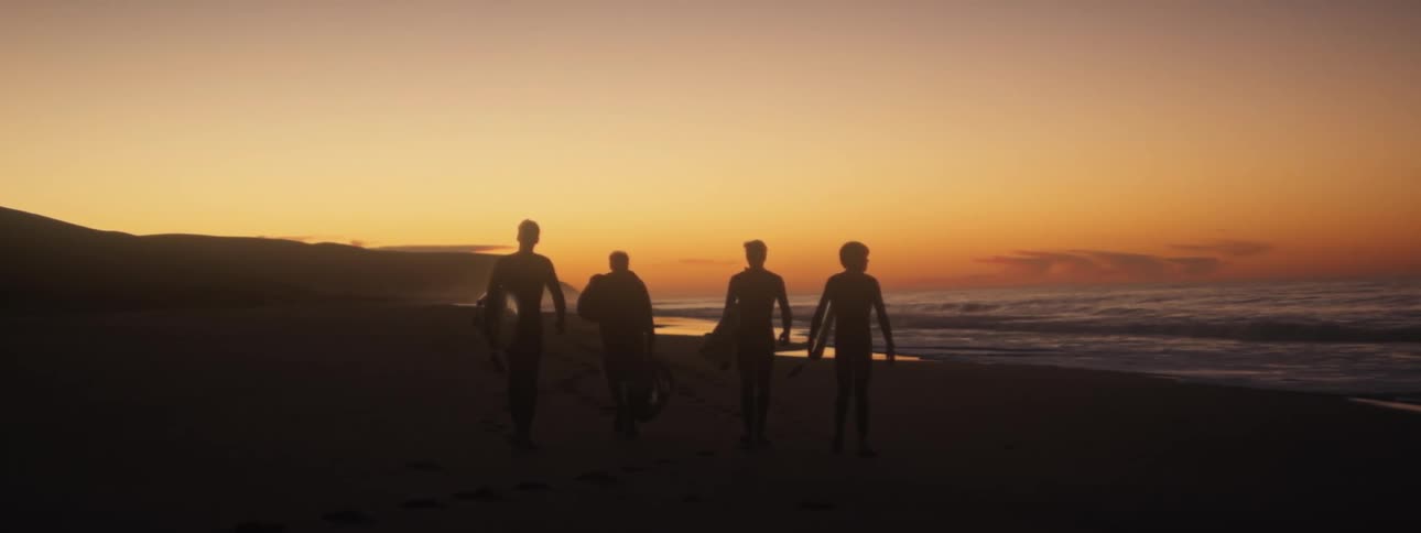 Four men carrying surf boards walk along the beach as the sun sets