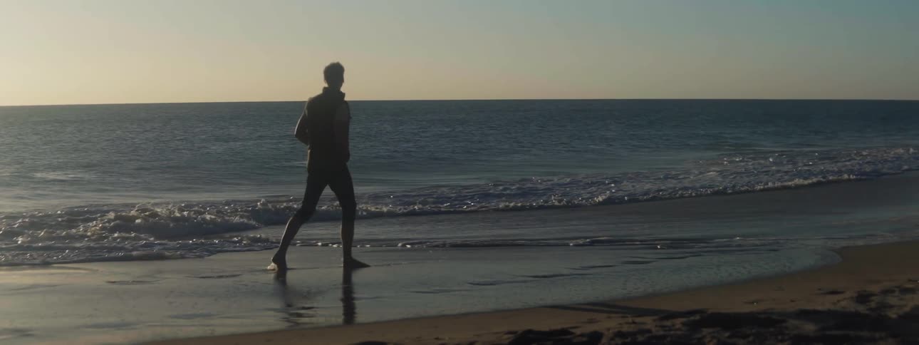 Man wearing coat and pants walks barefoot on the beach at sunset next to the sea