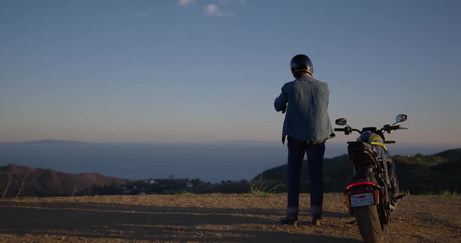 Man taking off his helmet while standing on the side of his motorcycle on top of a hill looking at the view ahead