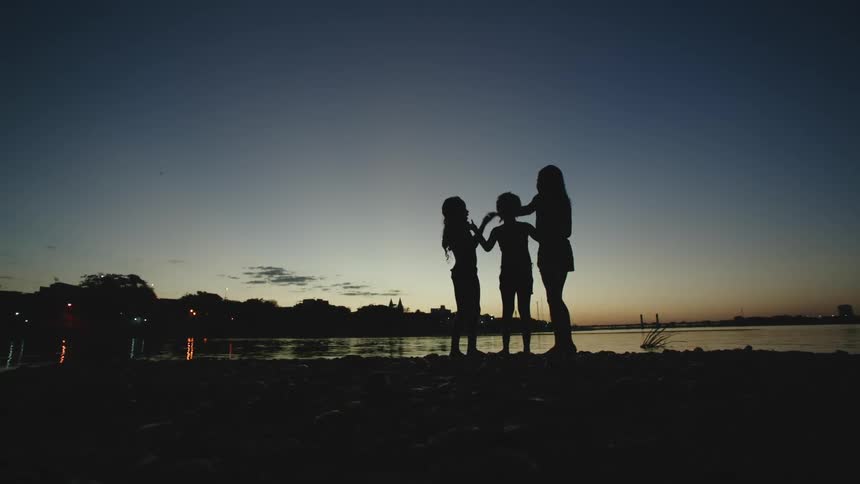 Three children stand on the shore and play hand games at dusk