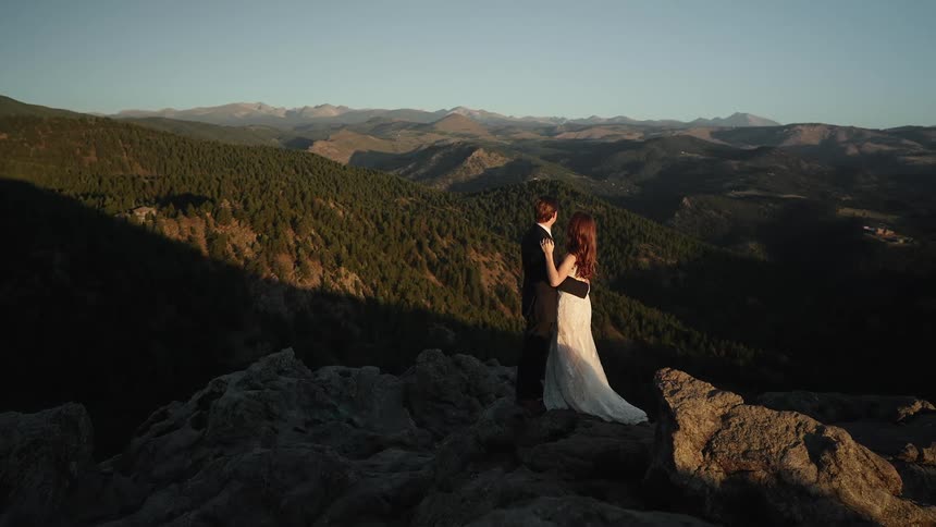 Bride and groom stand overlooking mountain range covered by forest