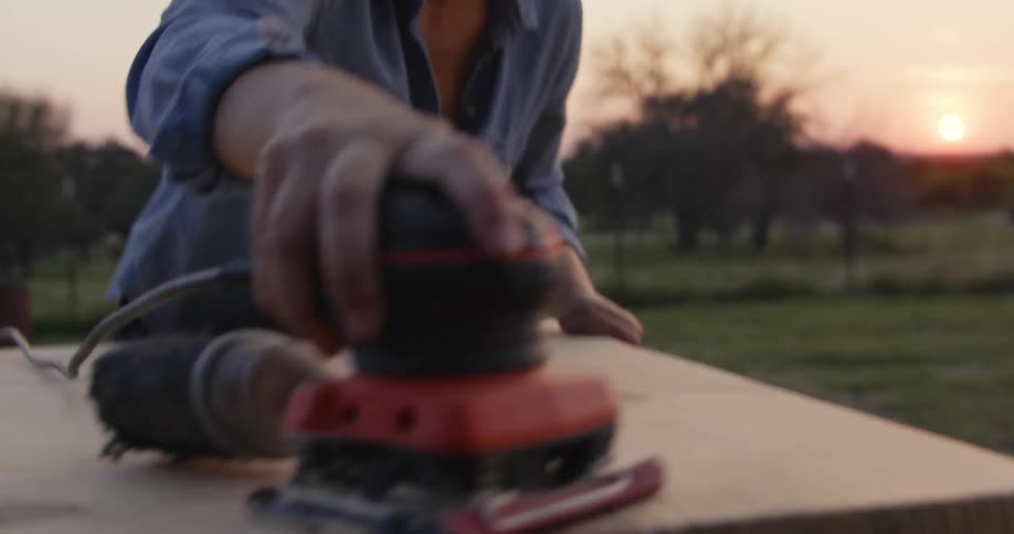 Woman uses a small power tool to sand a plank of wood in the countryside at sunse