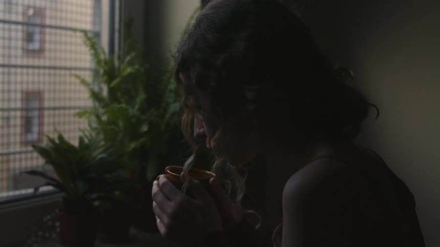 Woman with wavy hair stands next to houseplants and windows drinking from cup at apartment building