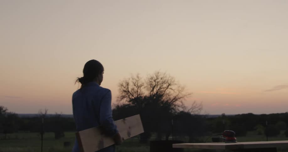 A woman carries a wooden plank over to a table she is building outside