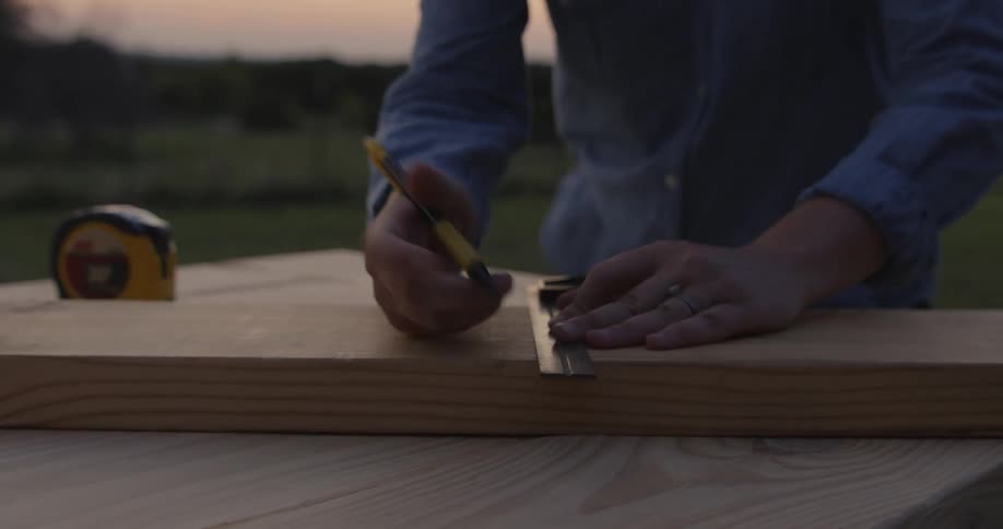 Woman uses a ruler to mark a line over a plank of wood over a workbench outdoors at evening