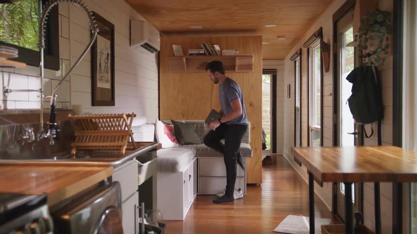 Young man carrying a laptop inside a small house sits on the couch near the kitchen