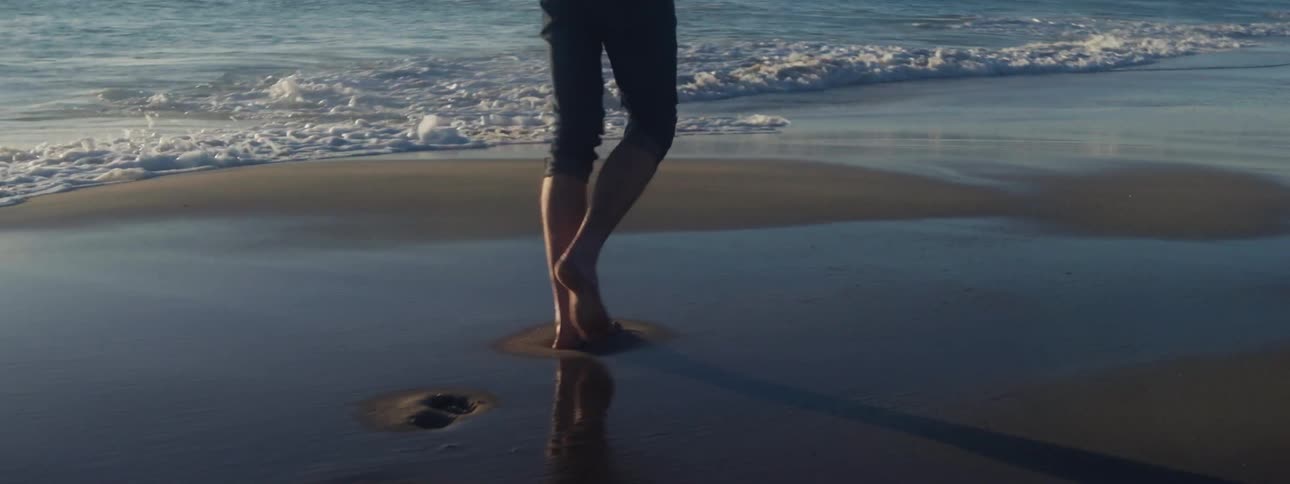 Close up of a person walking barefoot on the wet sand of a beach next to the ocean