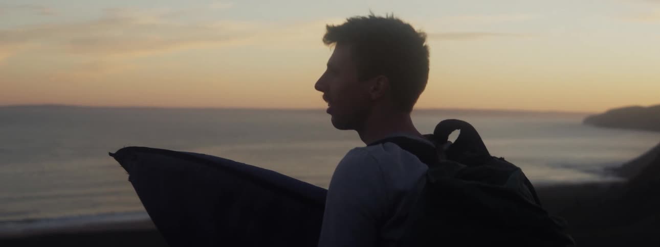 Young man holding a skateboard and wearing a backpack looks at the ocean at sunset