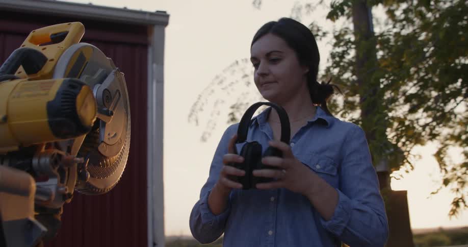A woman smiles as she puts on earmuffs before working on a circular saw set up near the barn