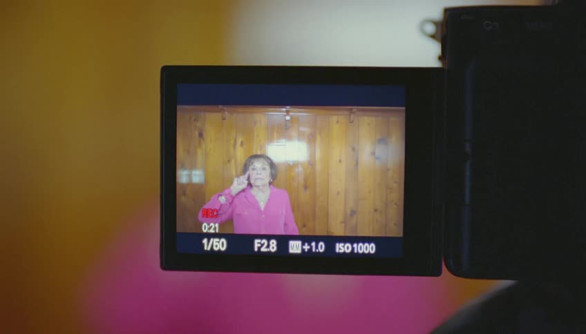Elderly woman stands in front of a video recorder and points at the camera