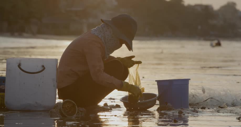 A person crouching over a wet landscape near a third world country town picking up things from the mud