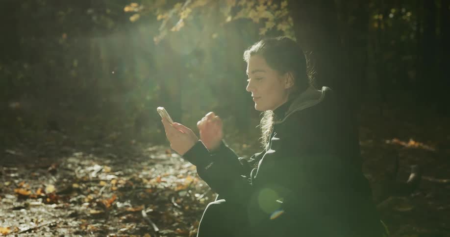 Young female cyclist sitting on the ground in the forest takes out her helmet and starts texting on her smartphone