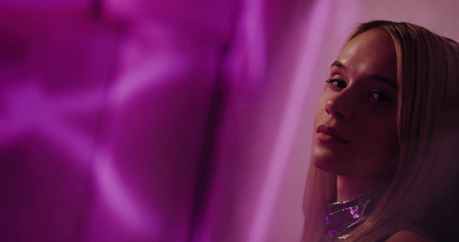A young woman stands under bright lights and turns her head as the air fills with fog