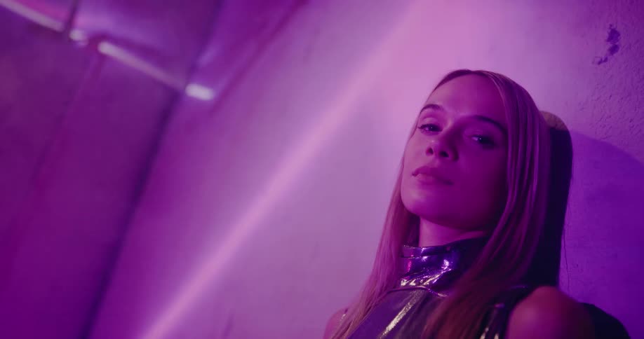 Blonde woman wearing metallic outfit leans against wall as colored lights flow over her