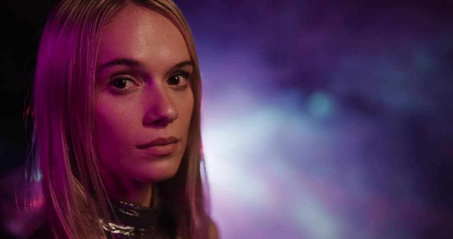 Pink light is shining on the face of a woman standing in a room filled with smoke and lights