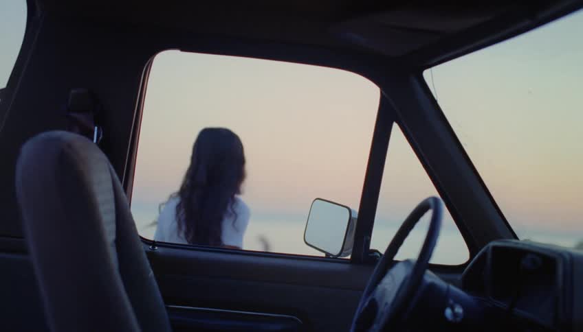 A teenage girl gets out of a truck and closes the door and walks towards the beach and looks out at the water