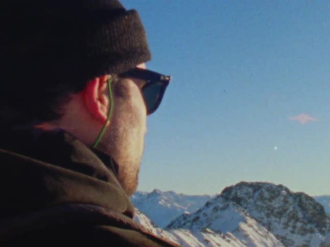 A man in a winter cap and sunglasses looking out over a snowy mountain range