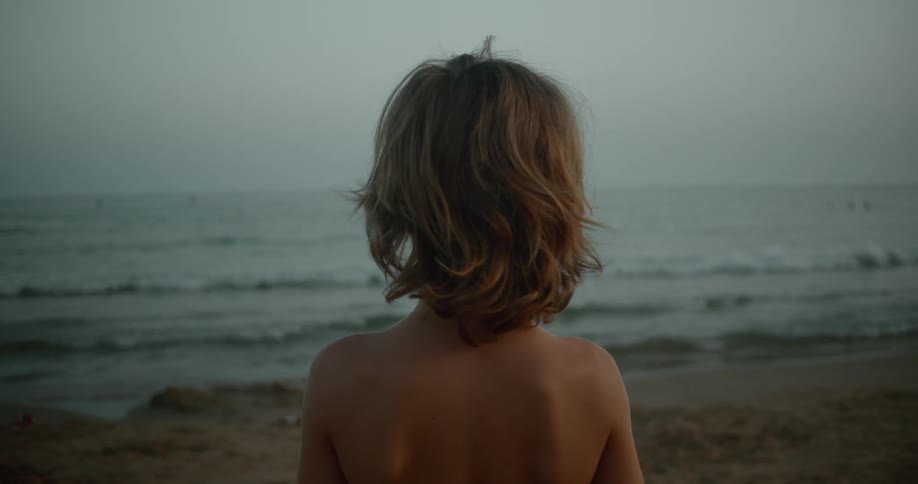 Back of a shirtless young blonde boy standing on the sand beach looking at the ocean at evening