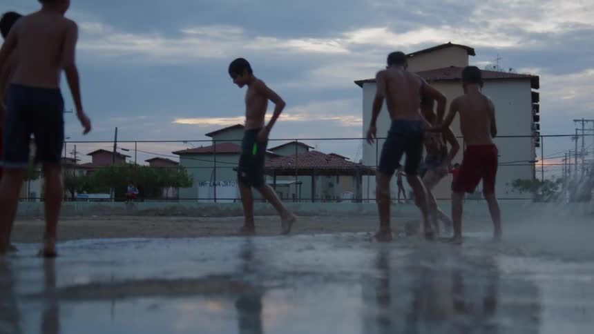 Group of young shirtless barefoot boys playing soccer on a park near a fountain at evening