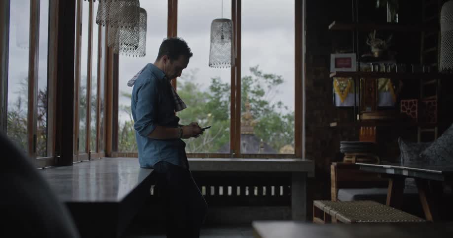Man leans back against table lining windows and texts on smartphone