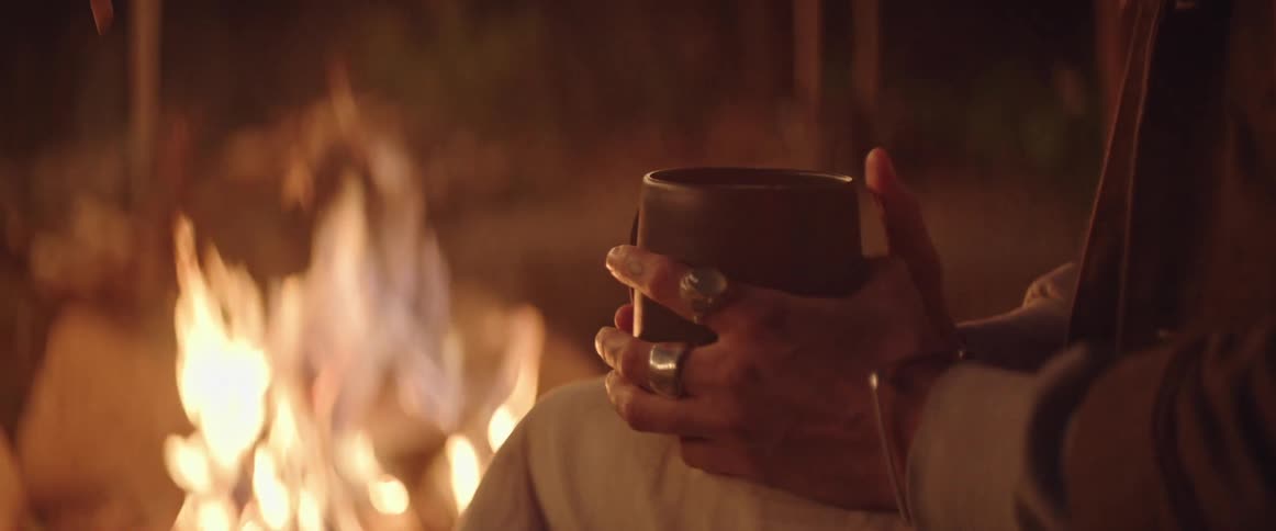 A person holding a hot beverage sits in front of a campfire at night