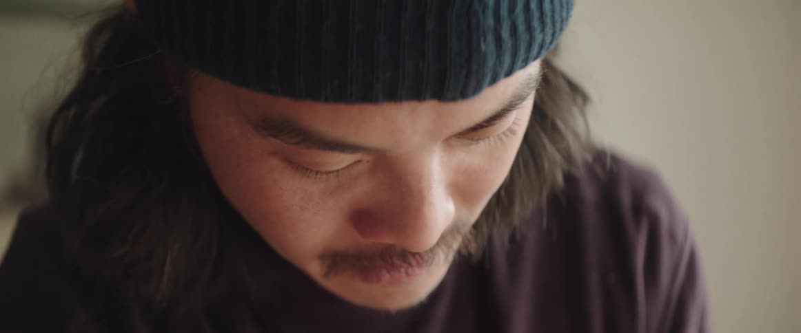 Close up of the face of a man with long hair and mustache working while wearing a beanie indoors