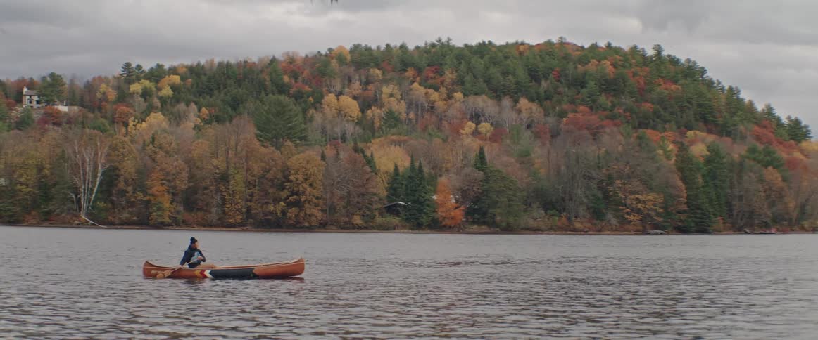 A person paddles a canoe across a lake on a cloudy autumn day