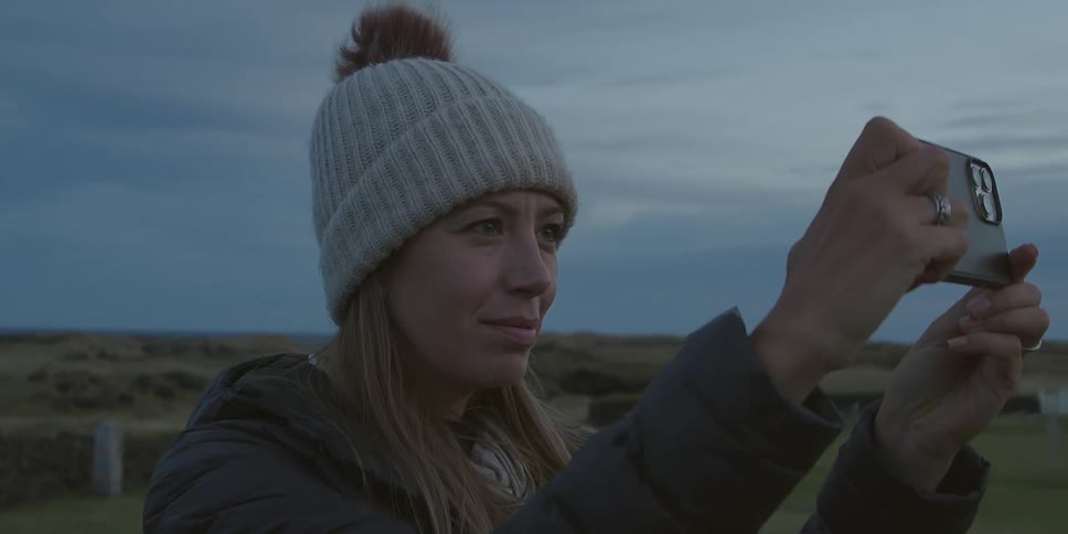 A woman wearing warm clothing holds up her phone to take a picture while outdoors