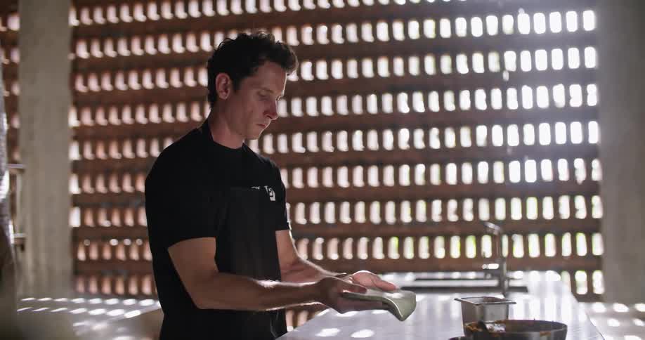 Chef works with dough at counter in restaurant kitchen lit by sunlight shining through openings designed into wall