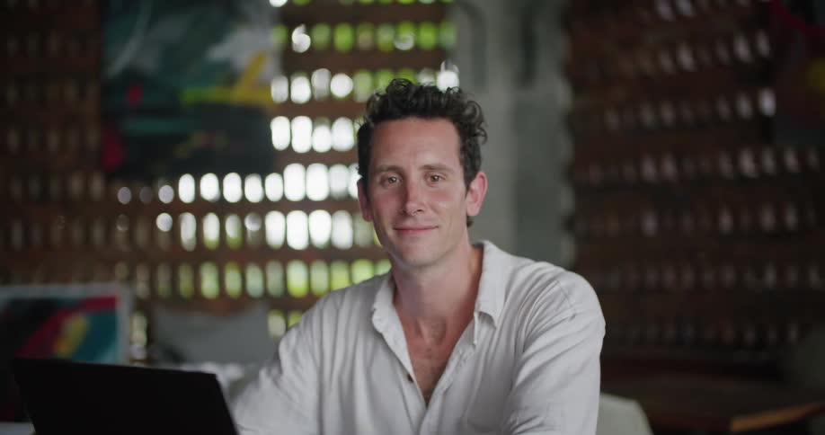 A man is sitting at a table behind a laptop computer and he looks up and smiles