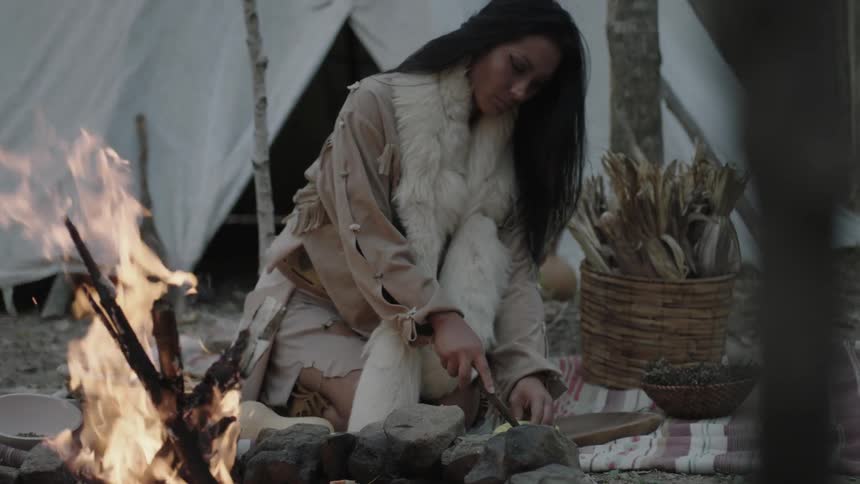 A native american woman carves something near a fire