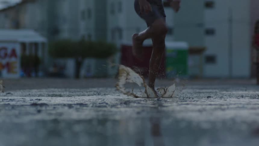 Group of barefoot boys splashing on a large puddle in front of residential buildings at evening