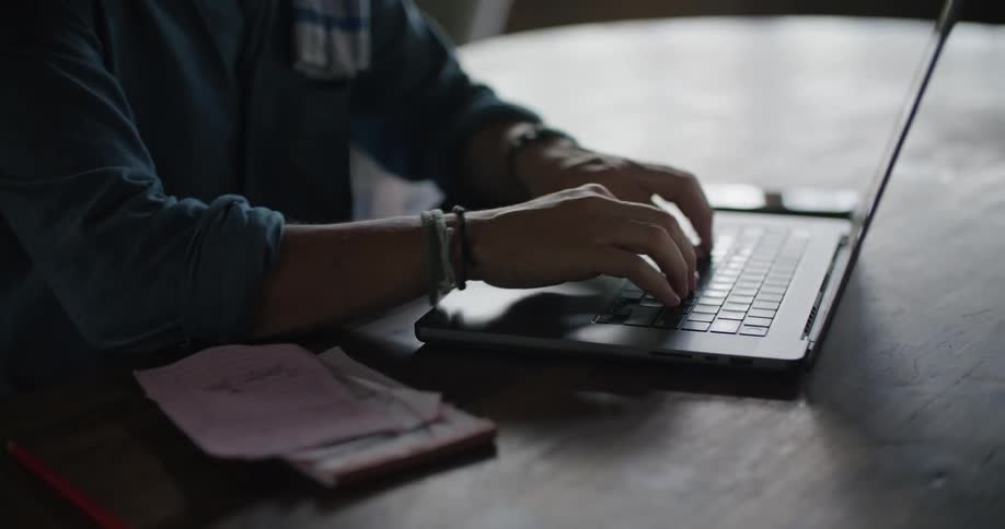A man is sitting at a table typing on a laptop computer and looking at papers on the table