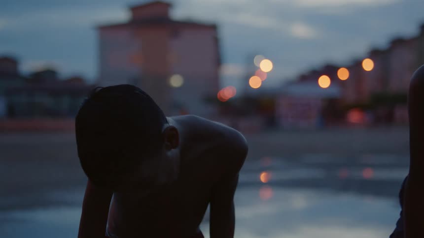 Shirtless boy laughing and gesturing in front of residential buildings at evening
