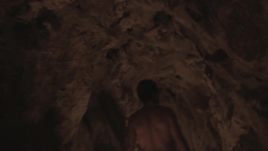 A shirtless man walks through a dark cave