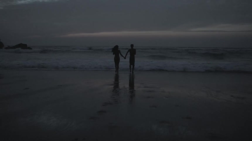 A man and woman holding hands run into the waves on a beach at dusk