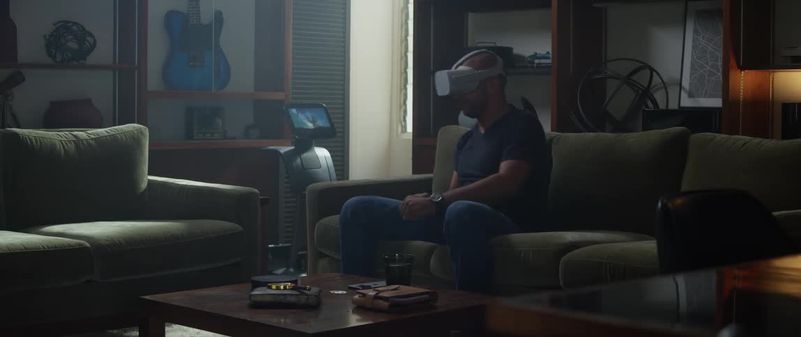A man is sitting on a living room couch wearing a virtual reality headset and moving his arm with a remote controller and shaking his finger as he plays