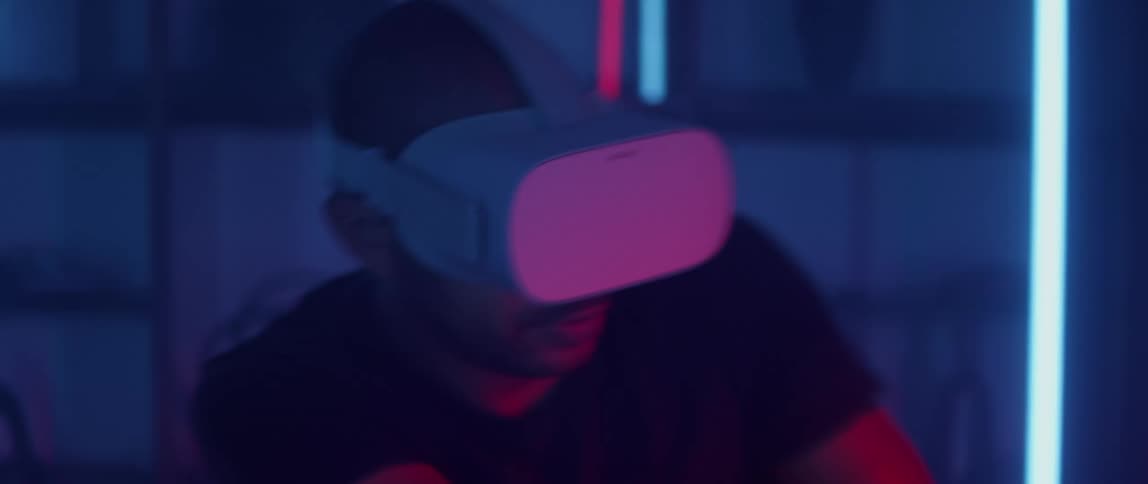 Neon lights reflect off a mans virtual reality headset as he jumps back and forth playing a game