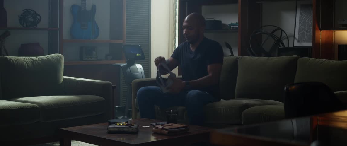 A man walks into the living room holding his phone and sits down on the couch and sets his phone on the table and puts on a virtual reality headset that is sitting on the table