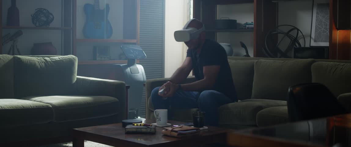 A man wearing a virtual reality headset sits on his couch and aims his controller