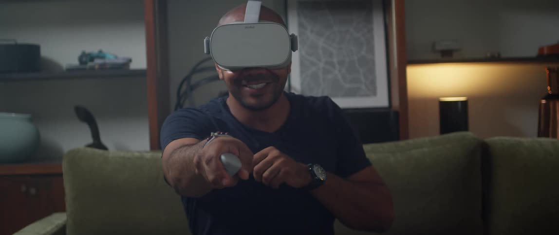 A man is sitting on a living room couch wearing a virtual reality headset and wildly moving his arm with a remote controller as he plays