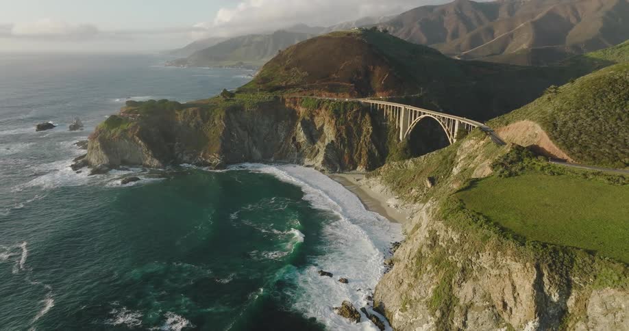 An aerial view of ocean waves flowing towards a shoreline with an arch bridge