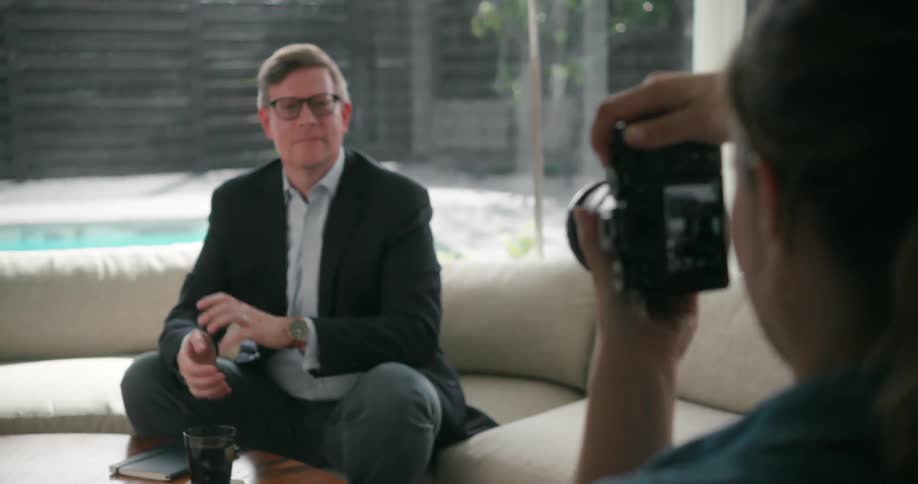 Female photographer takes photo of man wearing glasses blazer jacket and gray pants sitting on beige couch in the living room