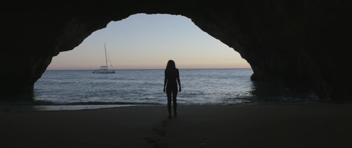 A woman walking under a rock formation like bridge, in the sand to the edge of the water